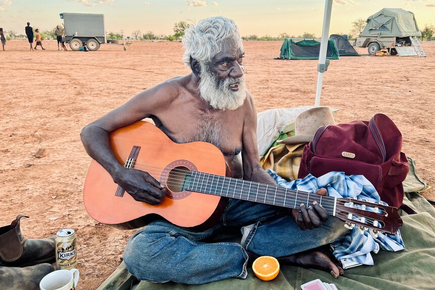 An elderly man with a white beard sits on the ground strumming a guitar