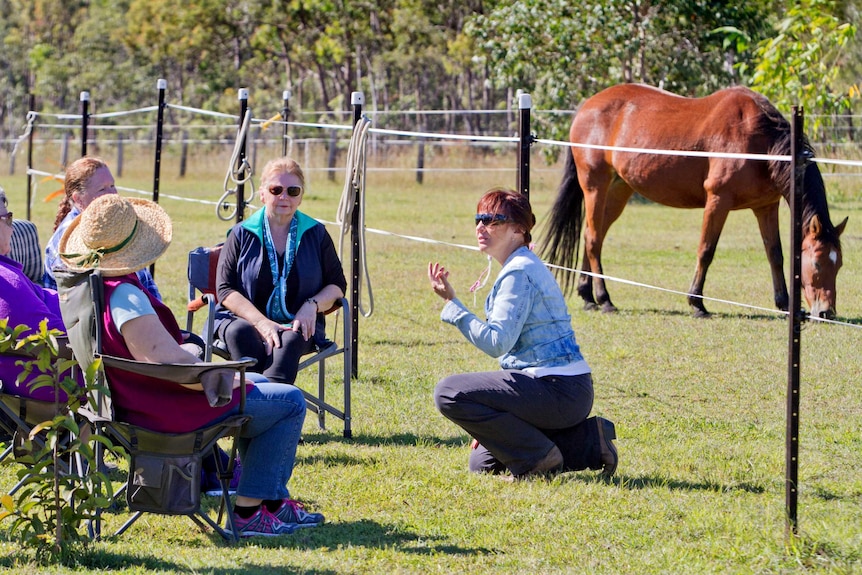 Helena Botros kneels as she speaks to four women in the horse paddock, she has red hair and wears sunglasses. Mid 30s.