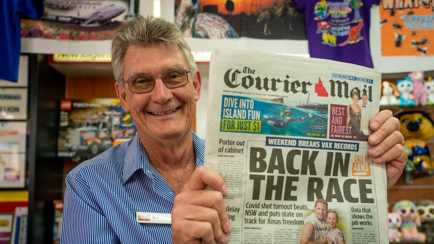 A smiling man stands behind a counter and holds up a copy of The Courier Mail with the headline "Back in the Race".