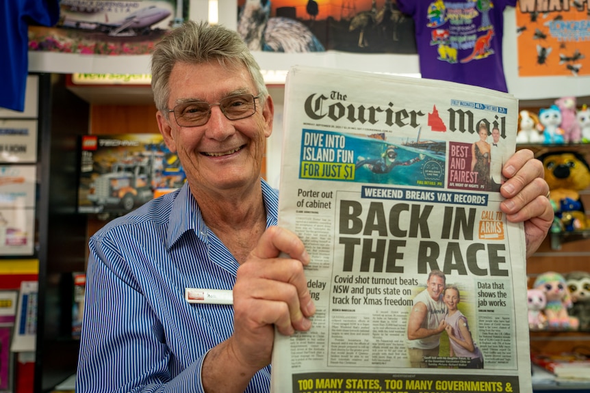 A smiling man stands behind a counter and holds up a copy of The Courier Mail with the headline "Back in the Race".