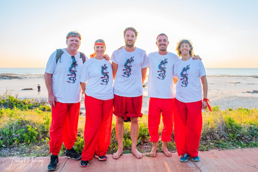 Five people stand in a row smiling in front of a beach