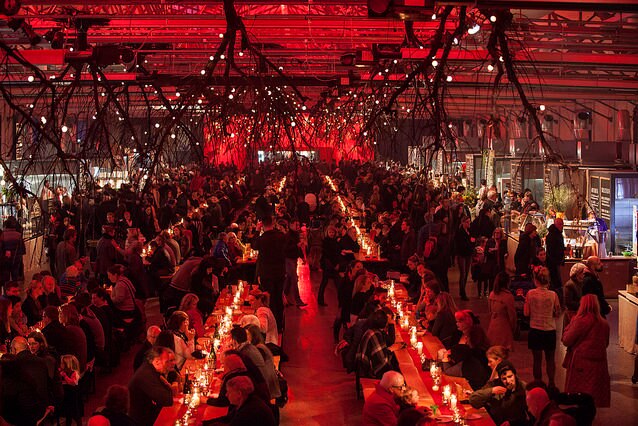 A large group of people gather in a dark dining hall under red lighting.