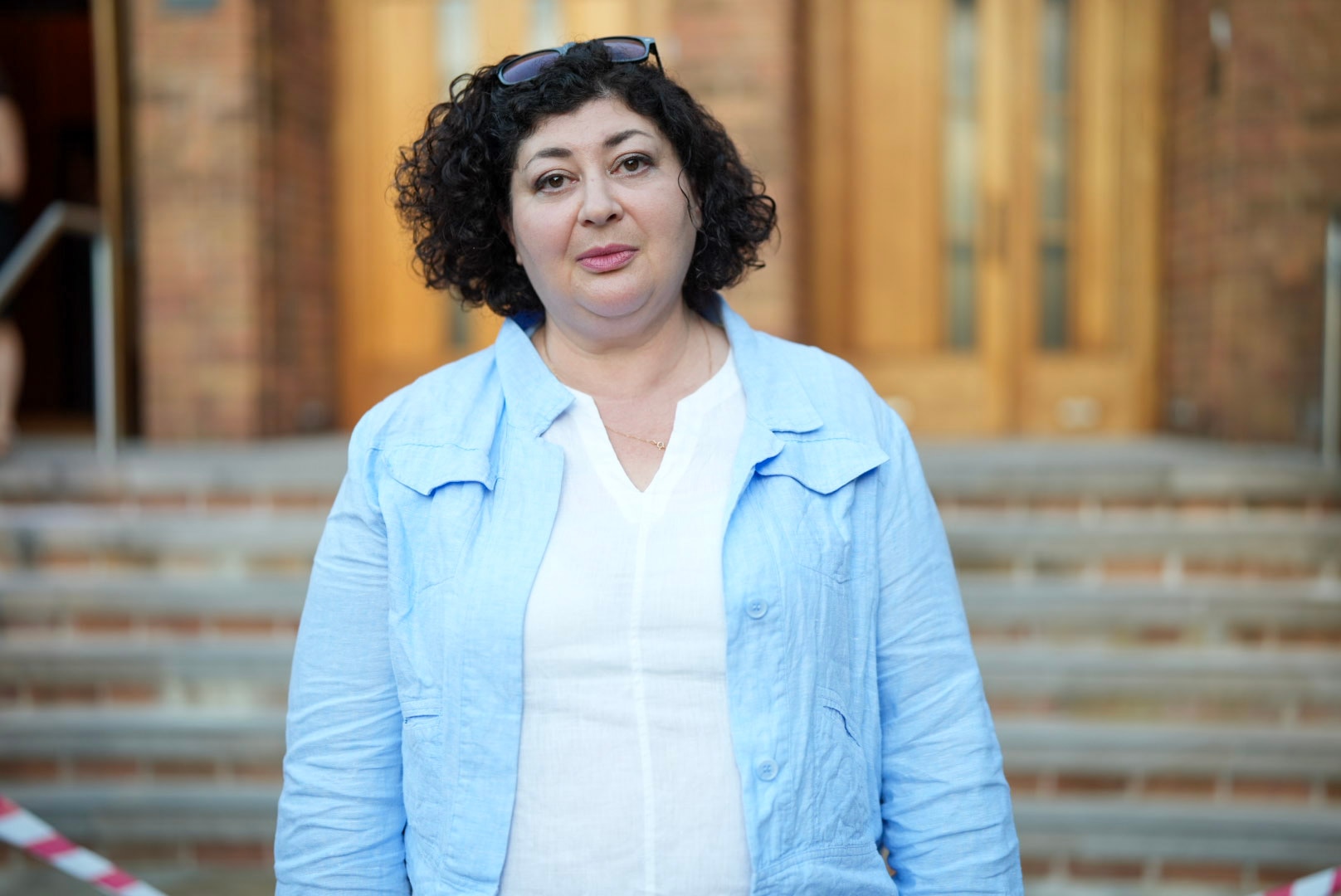 Uma mulher de jaqueta azul e blusa branca está em frente aos degraus de uma sinagoga.