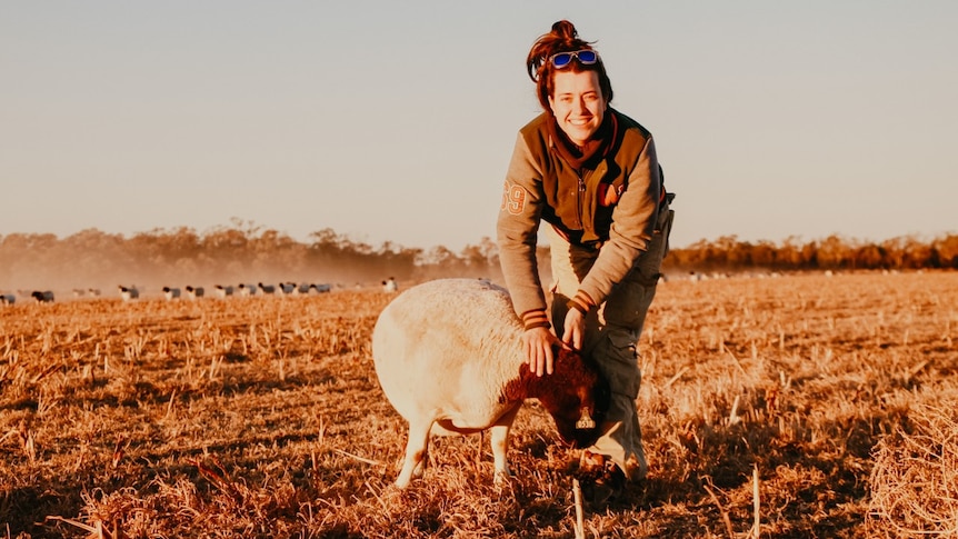 Female farm worker stands in a paddock smiling at camera whilst assessing a sheep