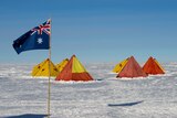 An Australian flag flies against a blue sky, in front of seven tents pitched on Antarctic ice.