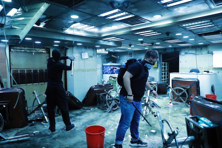 Protesters stand in a destroyed room full of glass and broken furniture holding sticks.