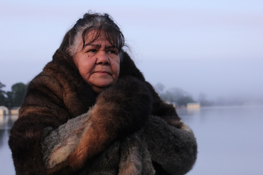 A woman stands in the early morning light, wrapped in a furry coat.