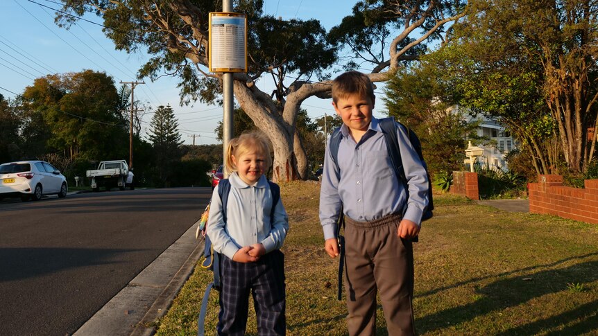 A little girl and boy in school uniform wait at the bus stop.