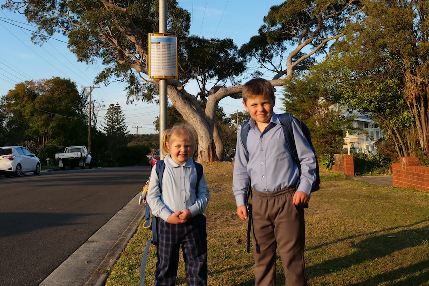 A little girl and boy in school uniform wait at the bus stop.