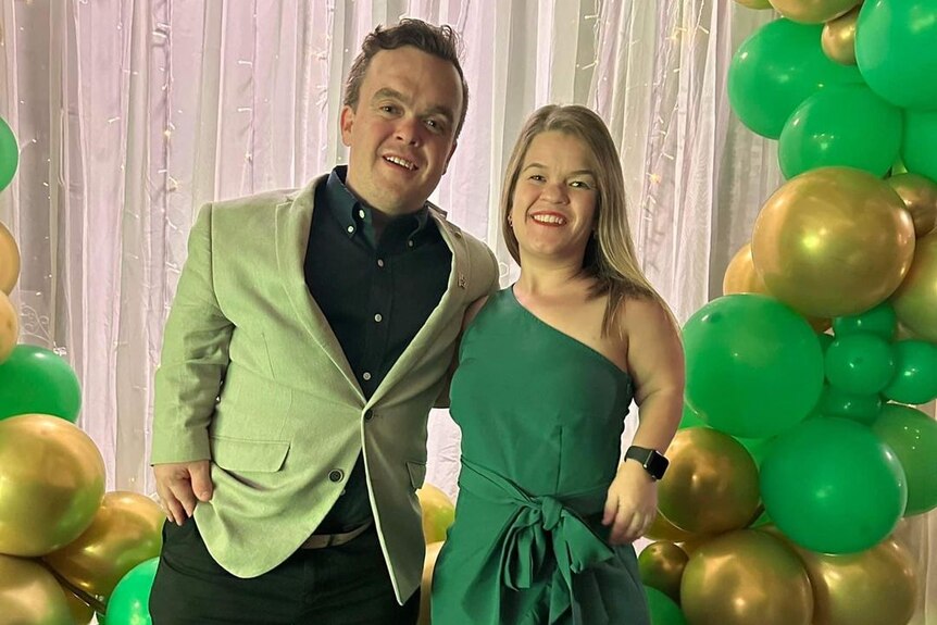 Michael and Sam, wearing green formal wear, are surrounded by green and gold balloons.