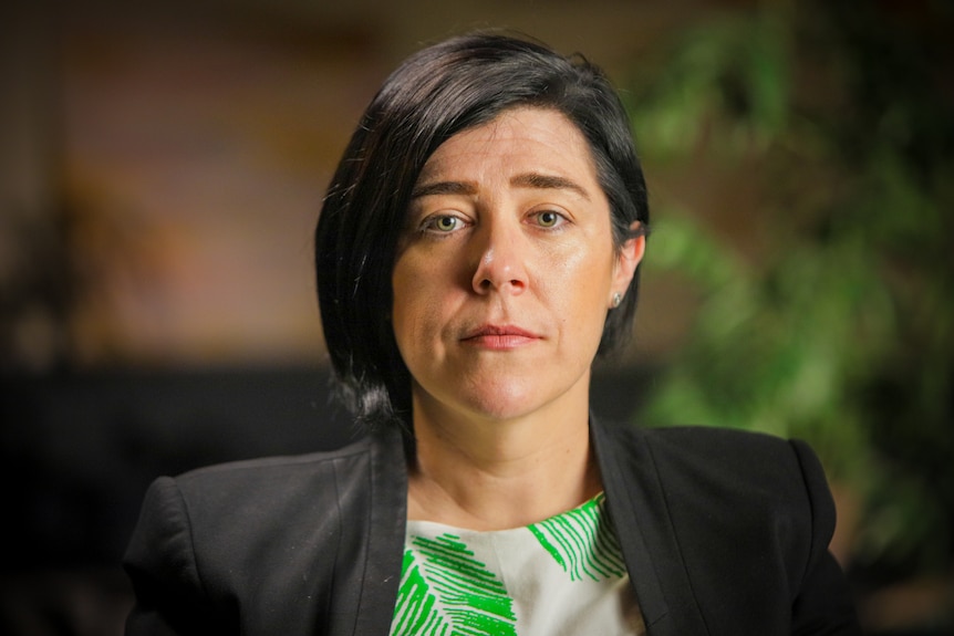 Woman wearing a white top with a green leaf print and a black blazer on top. She has short black hair.