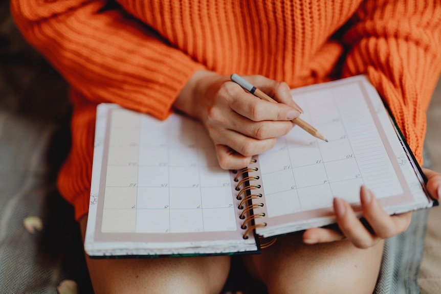 A woman wearing an orange sweater writes in a planner with a calendar.