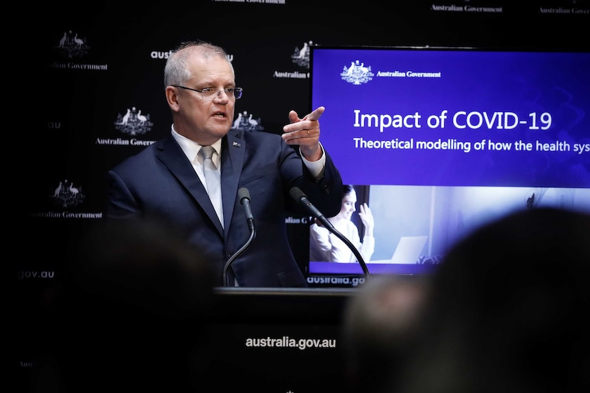 Scott Morrison stands in front of a screen with data about coronavirus. He points into the distance