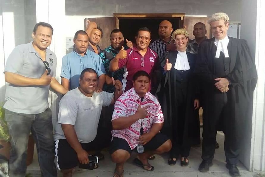 A group of people stand holding thumbs up next two a man and woman in black legal robes and wigs.