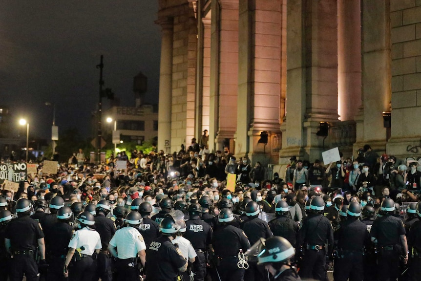 A large group of police wearing helmets, some holding clubs, surround a group of protesters near a large structure.