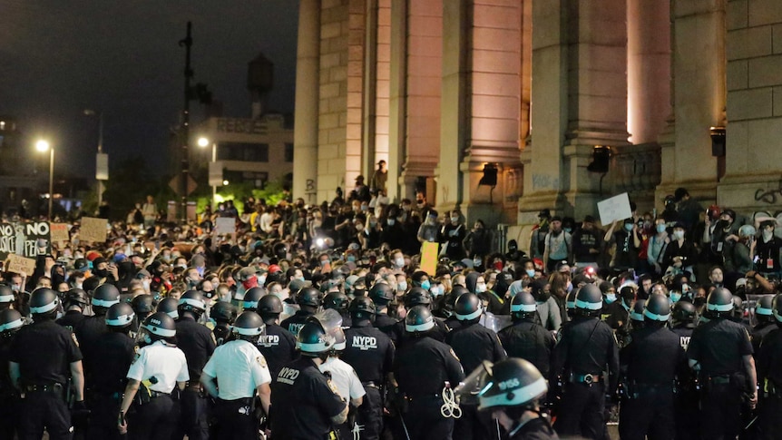 A large group of police wearing helmets, some holding clubs, surround a group of protesters near a large structure.