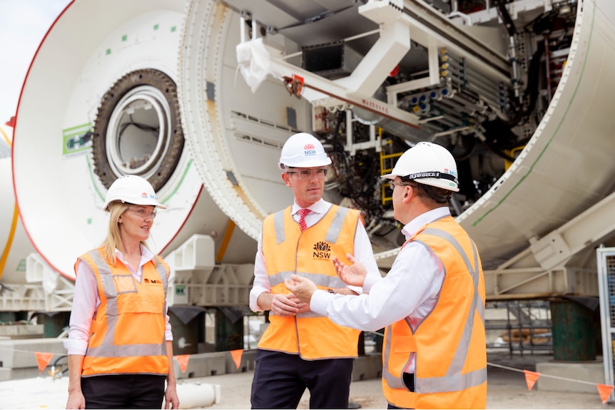 A woman, and two men, all with hard hats on, talking in frotn of a large machine