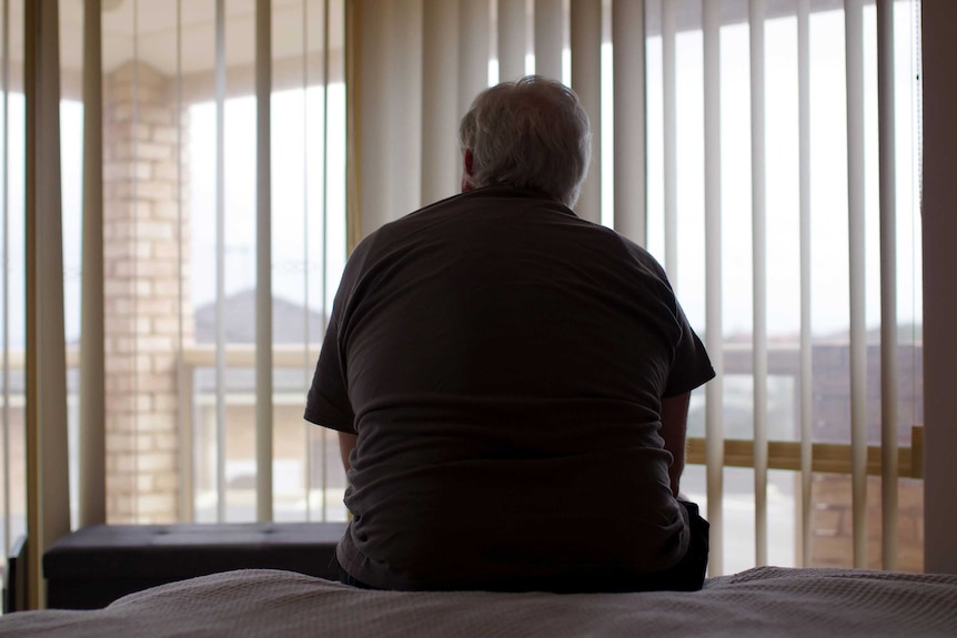 A man sits on a bed in a bedroom with his back turned looking out through windows.