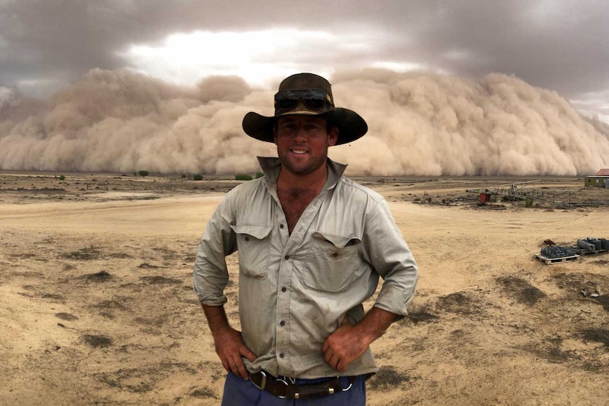 A farmer stands on a dusty property with his hands on his hips as a large dust storm rolls in and looms behind him.