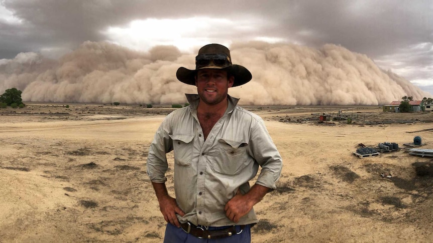 A farmer stands on a dusty property with his hands on his hips as a large dust storm rolls in and looms behind him.