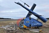 A crashed and snapped helicopter on the ground near a runway.