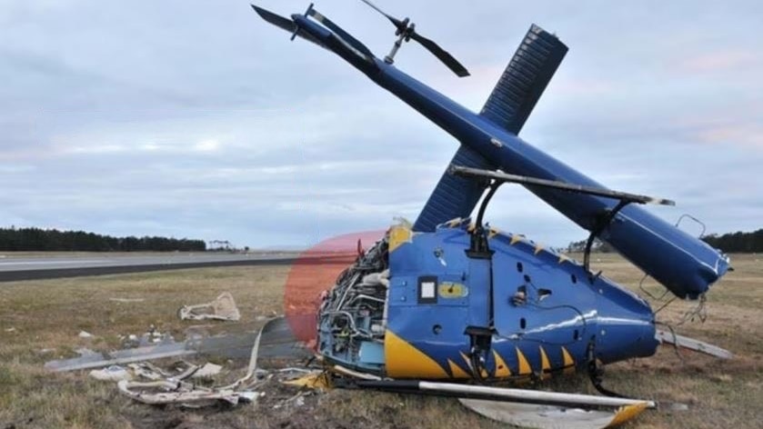 A crashed and snapped helicopter on the ground near a runway.
