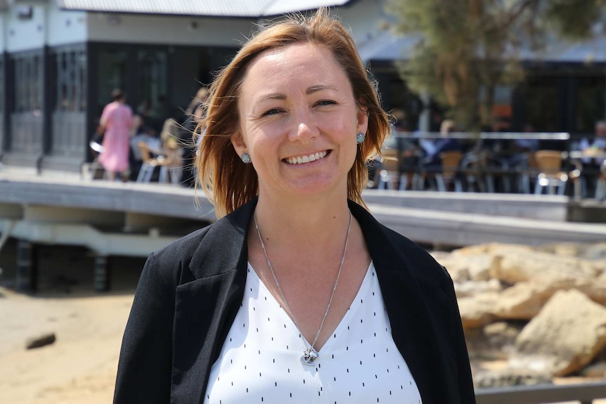 A head and shoulders shot of Coastal Waste Warriors organiser Kirstin Field standing on a beach in front of a building smiling.