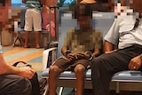 A young boy, whose face is blurred, sits in an airport with two men. He is wearing handcuffs.