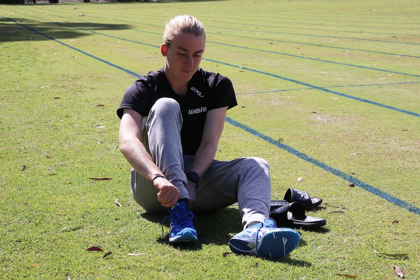 Declan Tingay puts his shoes on while sitting down on a grass field.