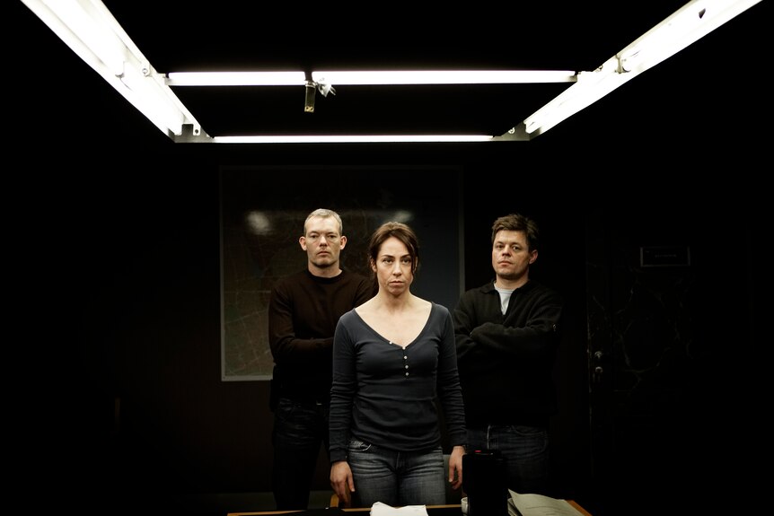 A woman and two men stand looking serious inside a police interrogation room