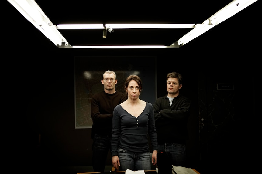A woman and two men stand looking serious inside a police interrogation room