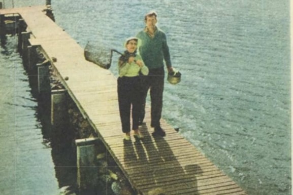 A woman and a man stand on a jetty, the woman is holding a fishing net.