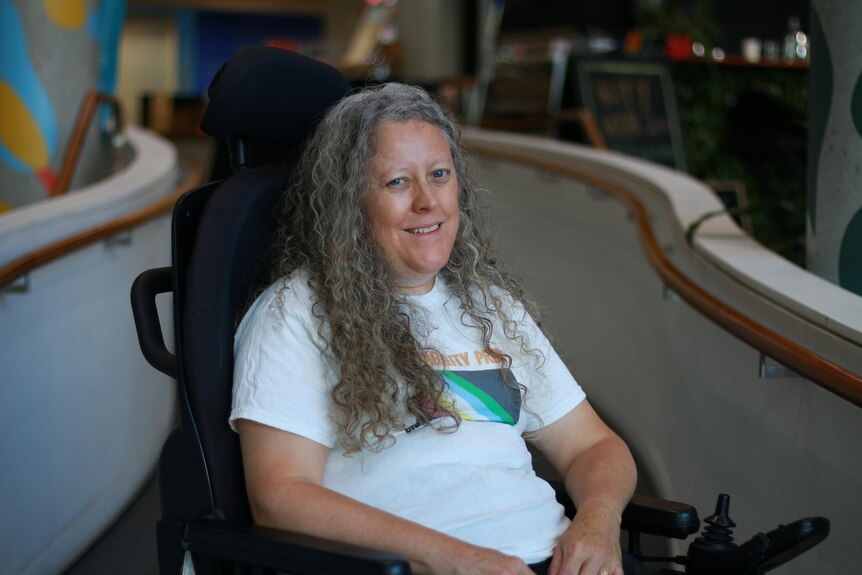A middle-aged woman sitting in a wheelchair. She is wearing a white t-shirt about disability pride