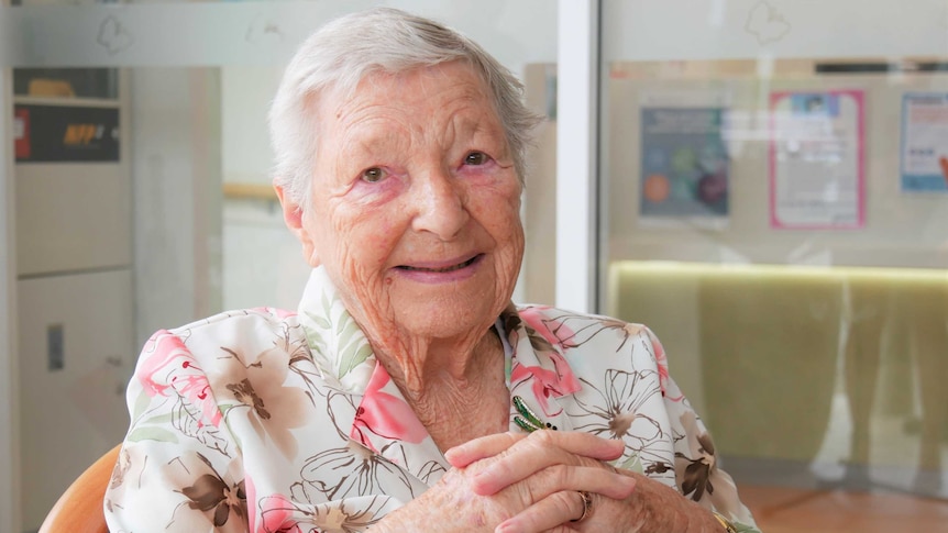 !00 year old woman smiling in her floral pink and white blouse, hands clasped, looking into the lense