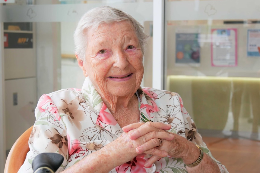 !00 year old woman smiling in her floral pink and white blouse, hands clasped, looking into the lense