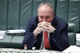 Joyce is sitting, elbows on desk, hands on face. He's wearing a dark suit and orange tie.