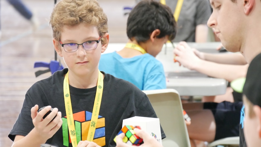 A boy sits at a table, holding a stopwatch as watches a man unscrambling a Rubik's Cube.
