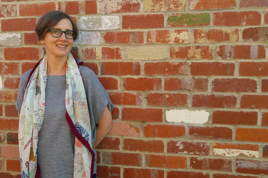 A woman with short hair, glasses, wearing a grey tshirt and scarf stands against a brick wall. 