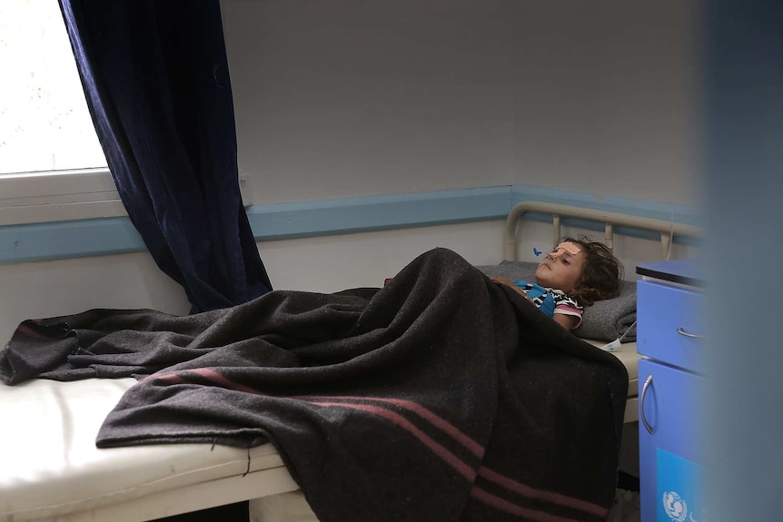 A young girl with a forlorn expression rests on a hospital bed under a blanket with two stripes.