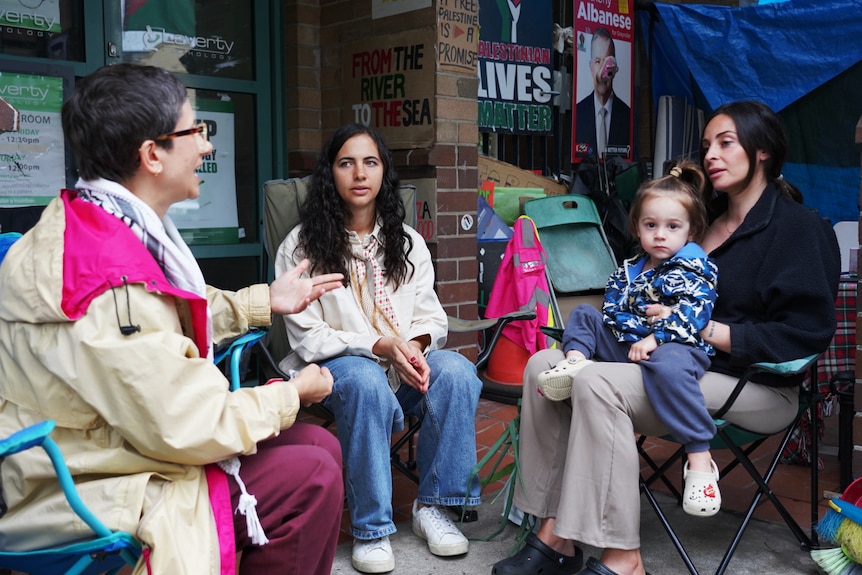 Three women and a child sitting on camp chairs talk to each other outside of an office bulding.