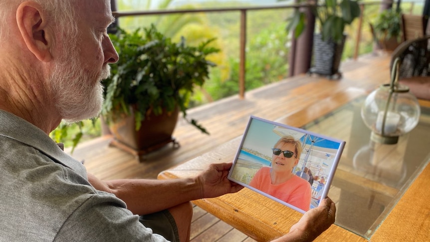 A man holds a photo of his wife, she is wearing a pink shirt and sunglasses in the framed photo. 