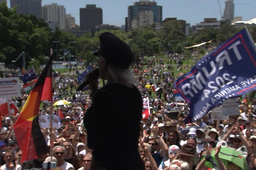 A speaker on stage addressing a large crowd, with a Trump flag and Aboriginal flag being waved behind them.