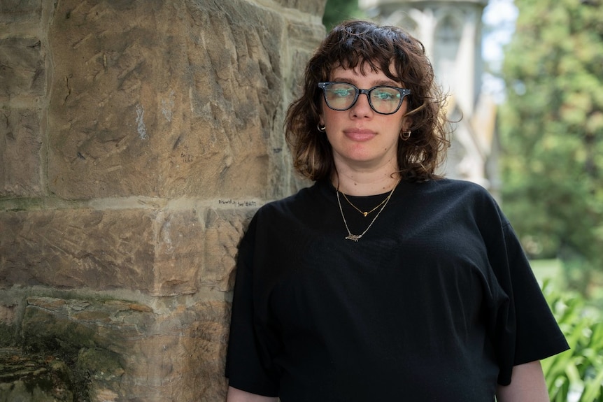 A young woman with large glasses stands next to a sandstone wall