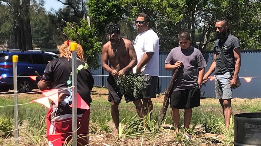 A group of people gather around a burial site marked with an Aboriginal flag.