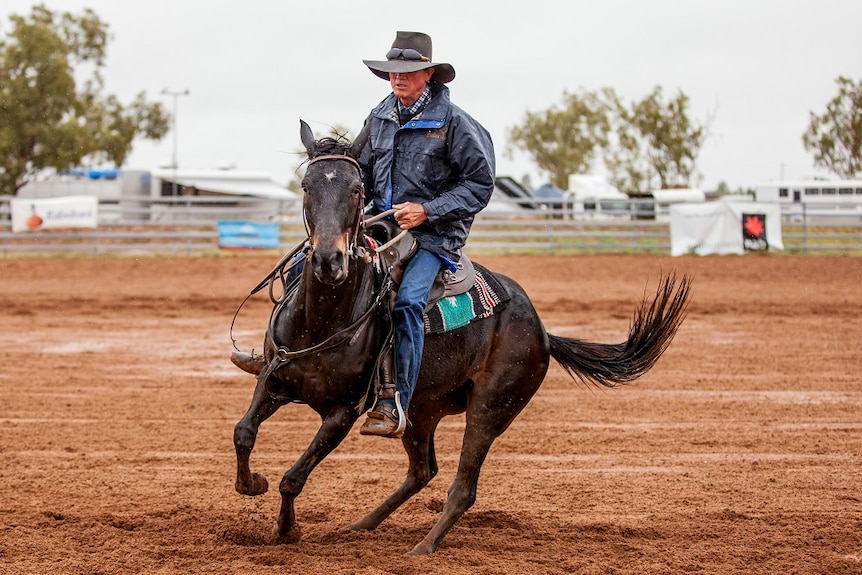 A man in waterproof clothing and large cowboy hat rides a dark horse in the rain