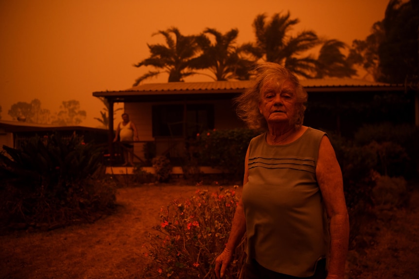 A elderly woman stands in front of a house with an ominous orange sky.