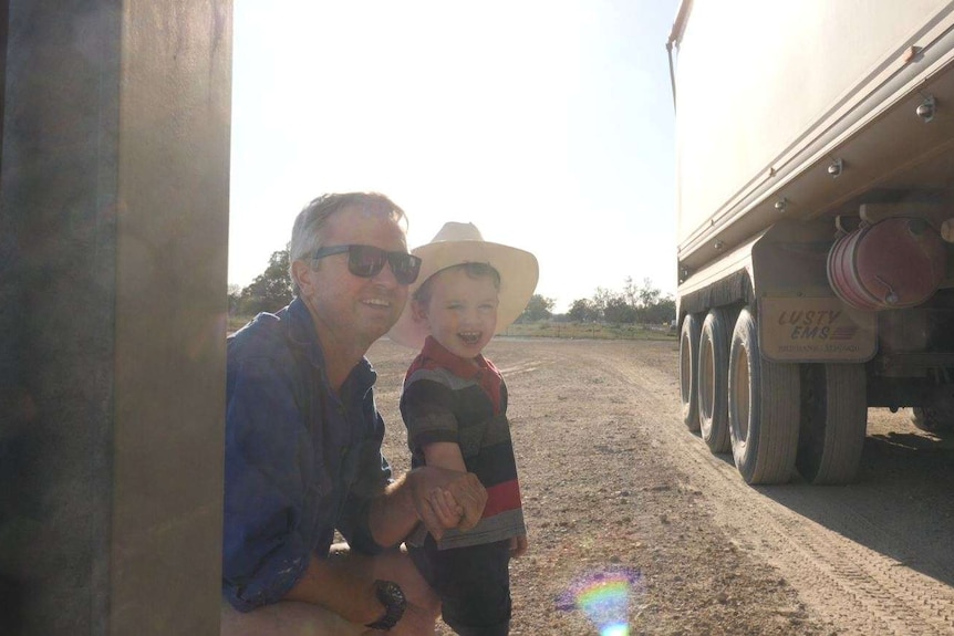 A man crouches beside of little boy in a cowboy hat beside a grain truck.