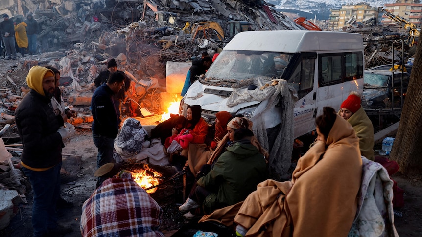 People sit around a fire next to rubble and damages near the site of a collapsed building in the aftermath of an earthquake