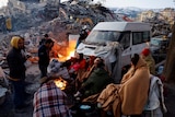 People sit around a fire next to rubble and damages near the site of a collapsed building in the aftermath of an earthquake