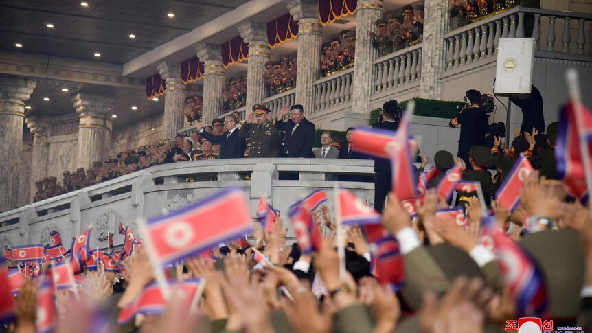Kim Jong Un and other dignitaries salue to flag waving crowd from balcony